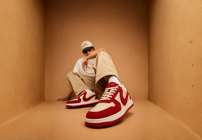 Person in a beige outfit crouching in a warm-lit set, wearing red and cream NAAW sneakers